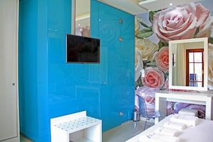 Una habitación con una pared de acento color turquesa, un televisor de pantalla plana montado sobre un banco blanco, un espejo con papel tapiz floral y una cama con toallas dobladas con cuidado.