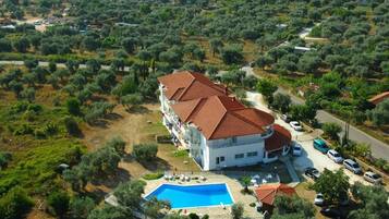 A large building with a swimming pool, surrounded by olive trees and a road.
