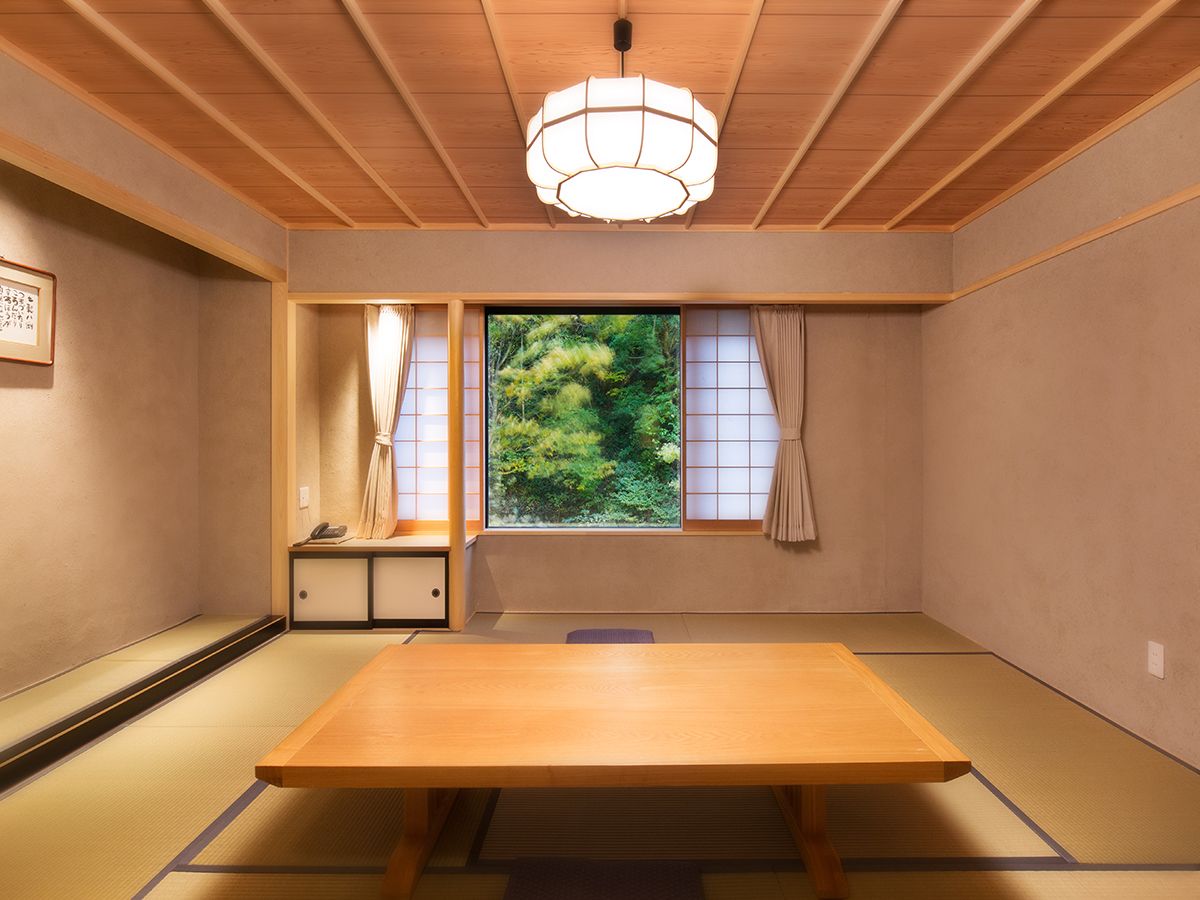 A traditional Japanese room with tatami flooring, a low table, sliding shoji doors, and a view of greenery outside.
