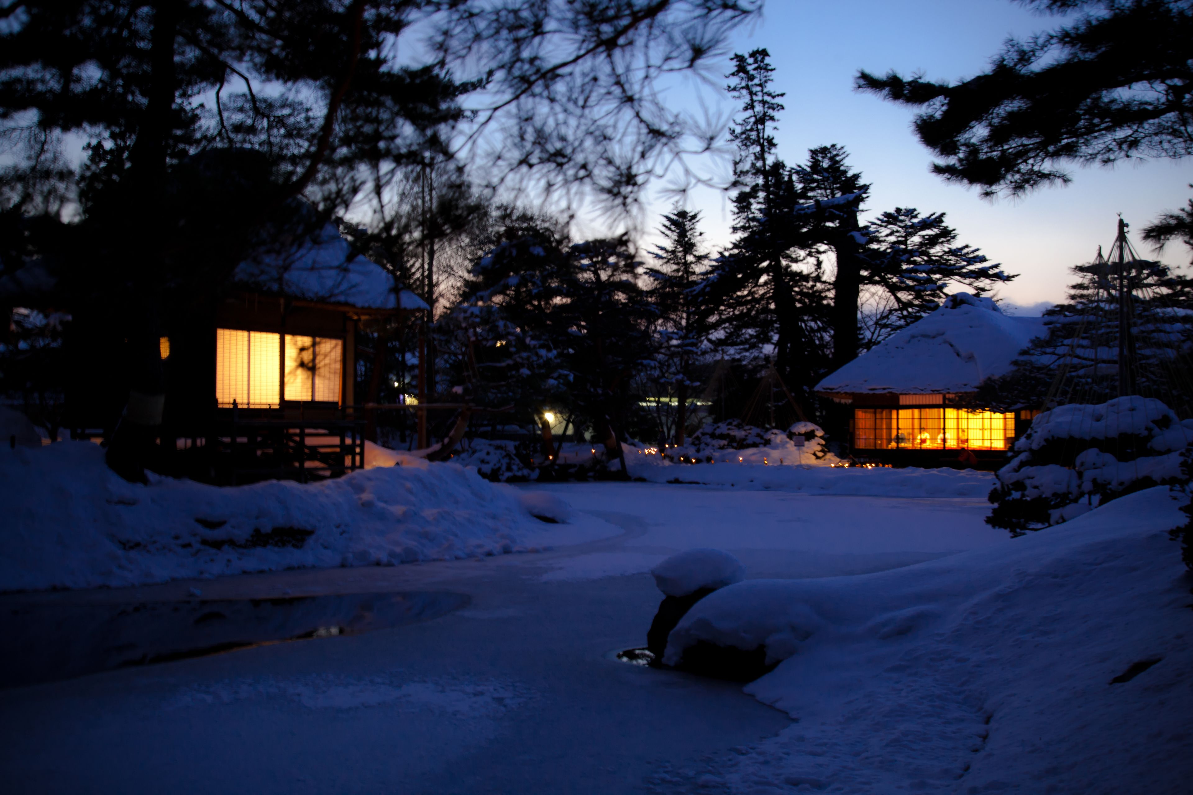 A snow-covered landscape with a wooden cabin and a building with illuminated windows.