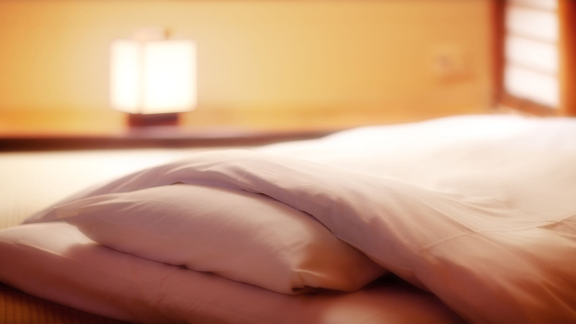 A bed with white bedding in a room with a warm, yellowish wall and a nightstand with a lamp.