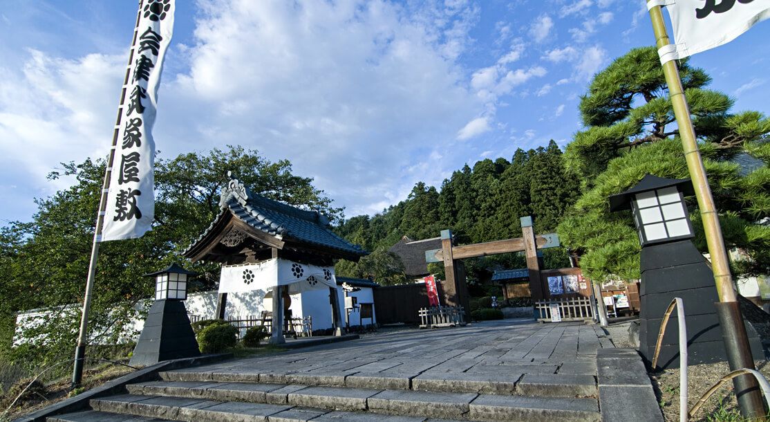 A traditional Japanese gate with a wooden structure and a tiled roof, surrounded by lush greenery and a clear sky.