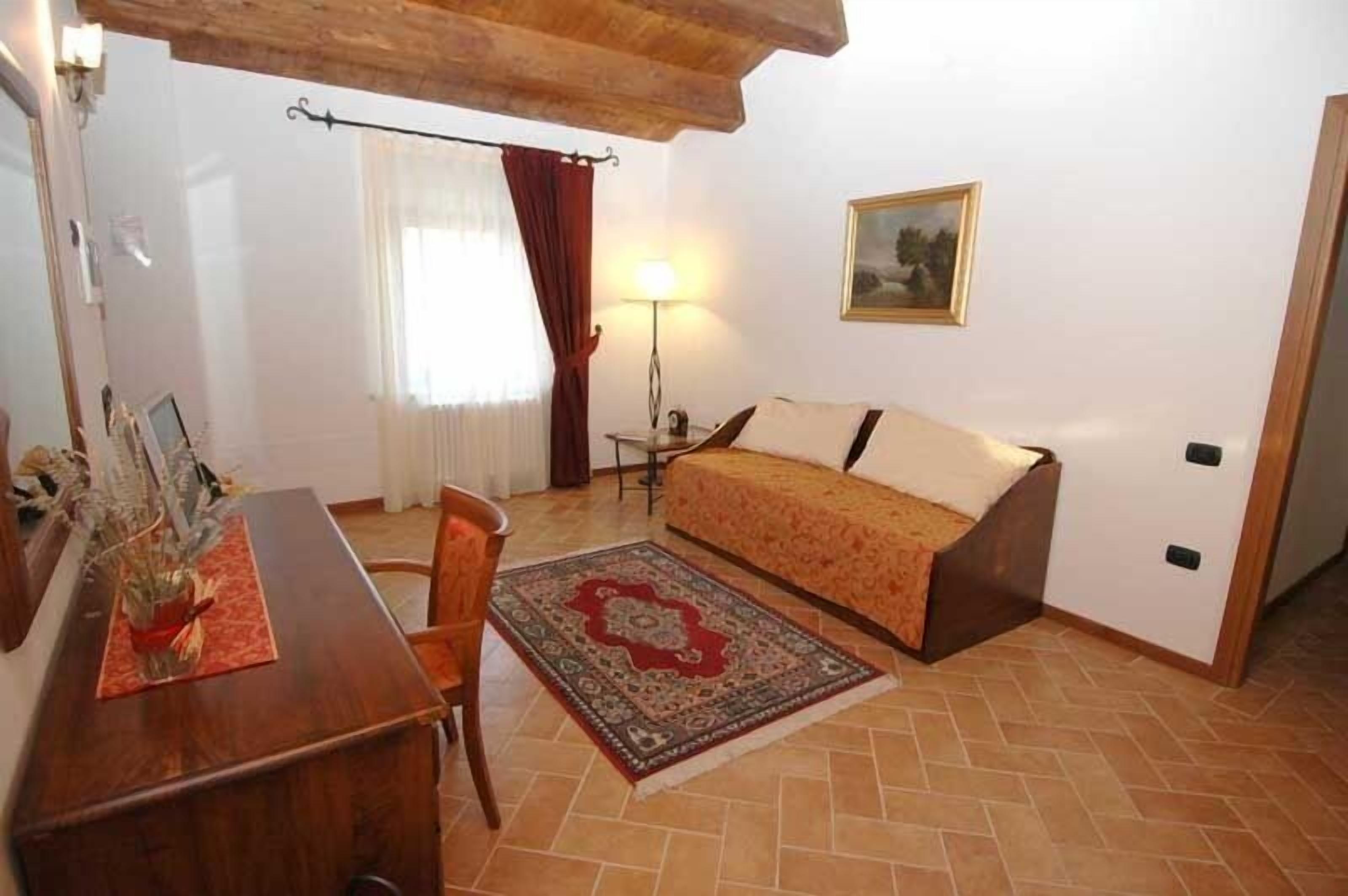 Quarto pequeno e aconchegante com uma cama, uma escrivaninha e uma cadeira. Há uma luminária em uma mesinha de cabeceira e um quadro na parede.