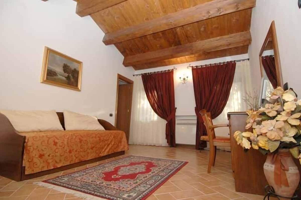 A cozy room with a sofa, a chair, a wooden ceiling, a painting, and a vase with flowers.