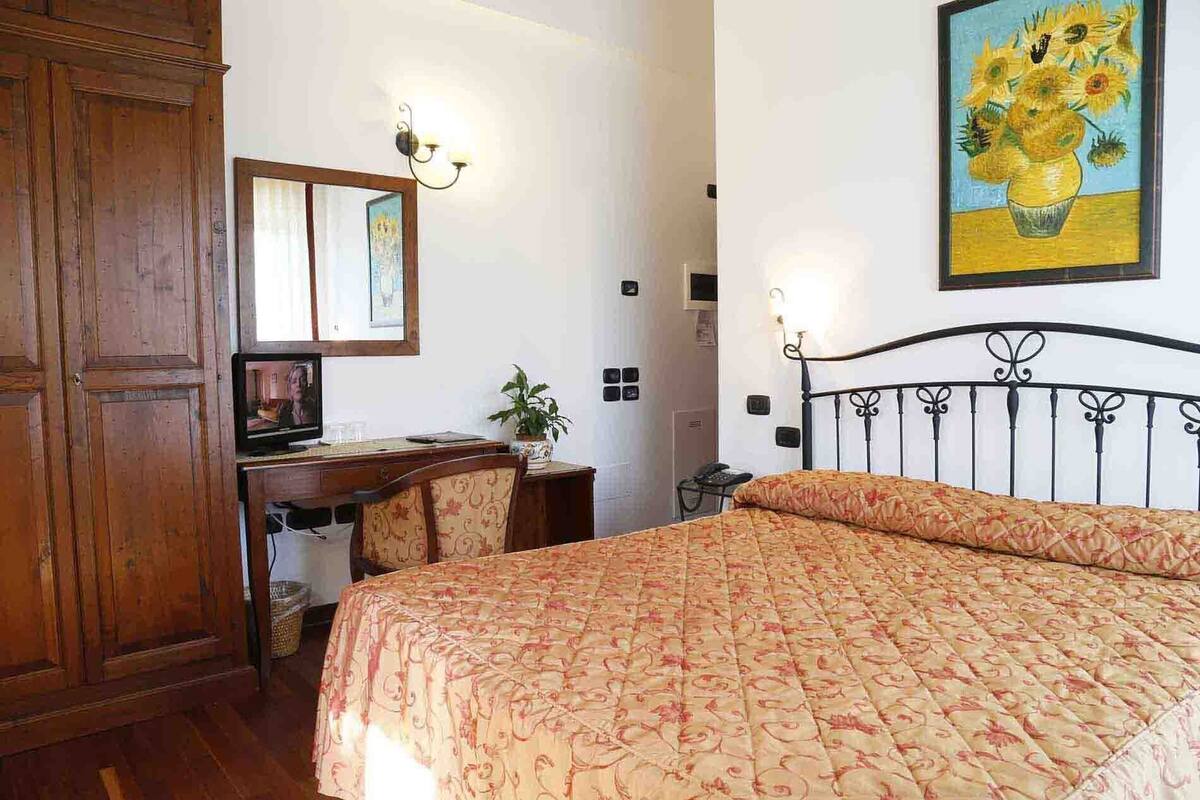 A hotel room with a bed, a desk with a chair, a wardrobe, and a painting on the wall.