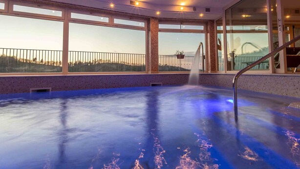 An indoor swimming pool with a view of a mountain range, large windows, and a staircase leading to a balcony. - Colleverde Country House Beauty&Wellness (Urbino)