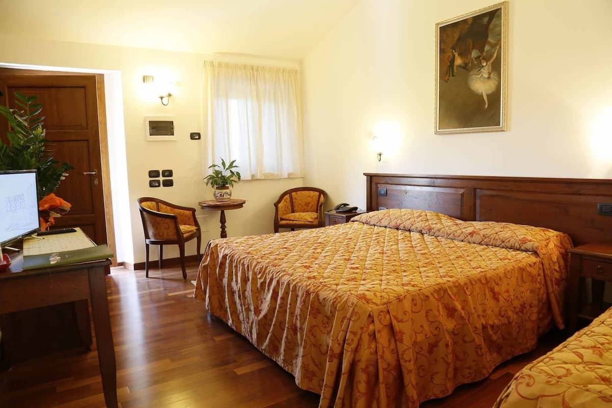 A hotel room with a bed, a desk with a computer, a chair, a small table with a plant, and a painting on the wall.