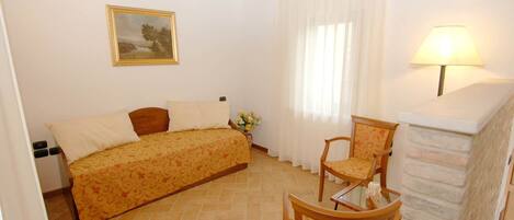 A bedroom with a bed, a chair, a lamp, a painting, and a window with curtains.