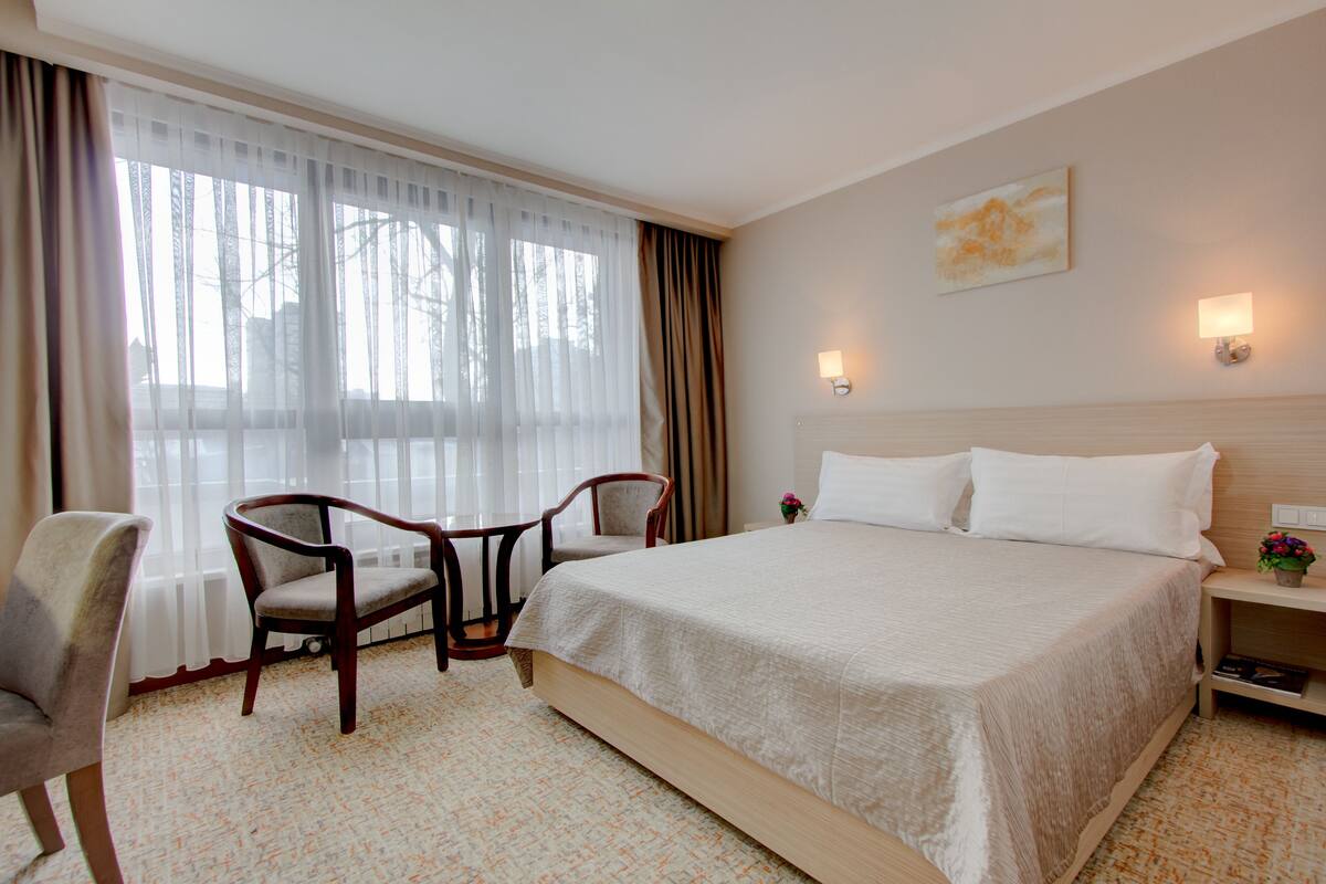 A hotel room with a large bed, two armchairs, a small table, a nightstand with a lamp, and a window with sheer curtains.