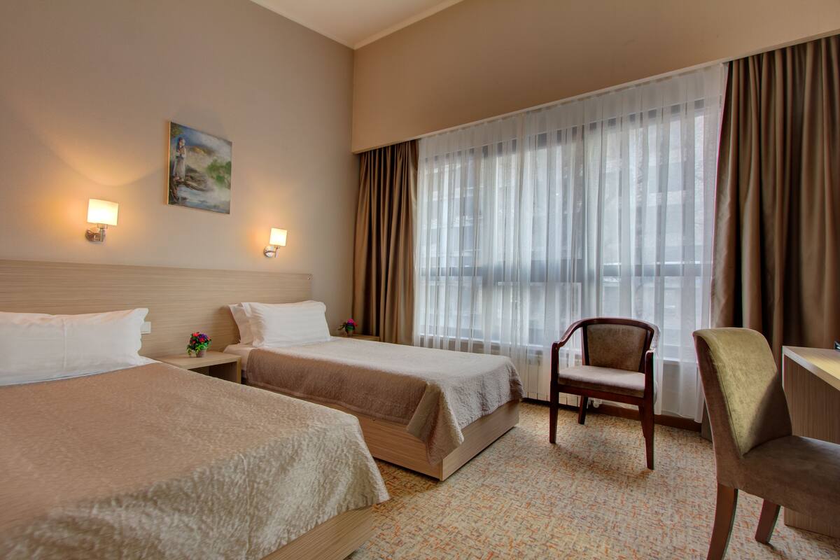 A hotel room with two beds, a desk, a chair, and a large window with curtains.