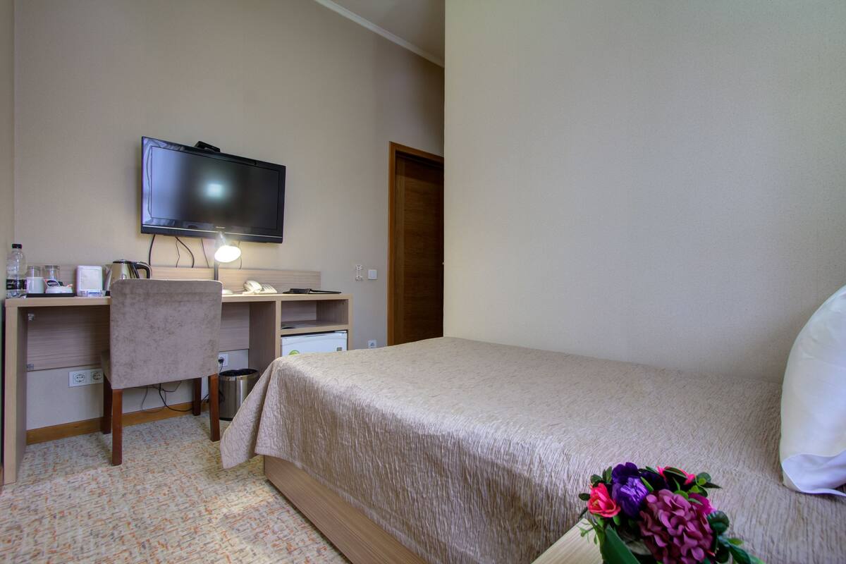 A hotel room with a bed, a desk with a computer, a television, and a small trash bin.