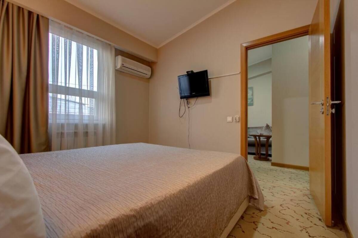A hotel room with a bed, an air conditioning unit, a television mounted on the wall, and a door leading to another room.