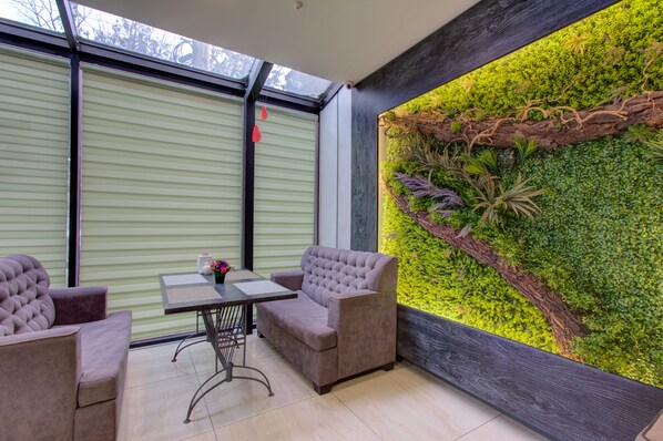 Una habitación con un ventanal amplio, un sofá acolchado, una mesita pequeña y una jardinera colgante con follaje verde.