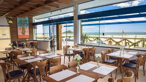 A restaurant with wooden tables and chairs, large windows with a view of the beach, and a painting on the wall. - Praia Hotel Imbituba (Imbituba)