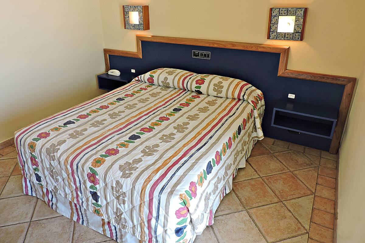 Una cama bien hecha con una colcha y cojines estampados con motivos florales, flanqueada por dos mesitas de noche con lámparas, en una habitación con suelo de baldosas y cabecera oscura.