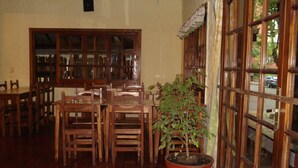 Comedor con mesas y sillas de madera, una planta en maceta y ventanas grandes con cortinas.