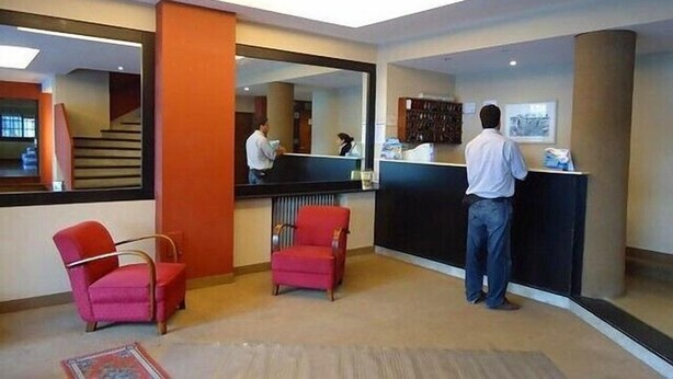 Zona de recepción de un hotel con mostrador, dos sillones rojos y un espejo grande que refleja el interior.