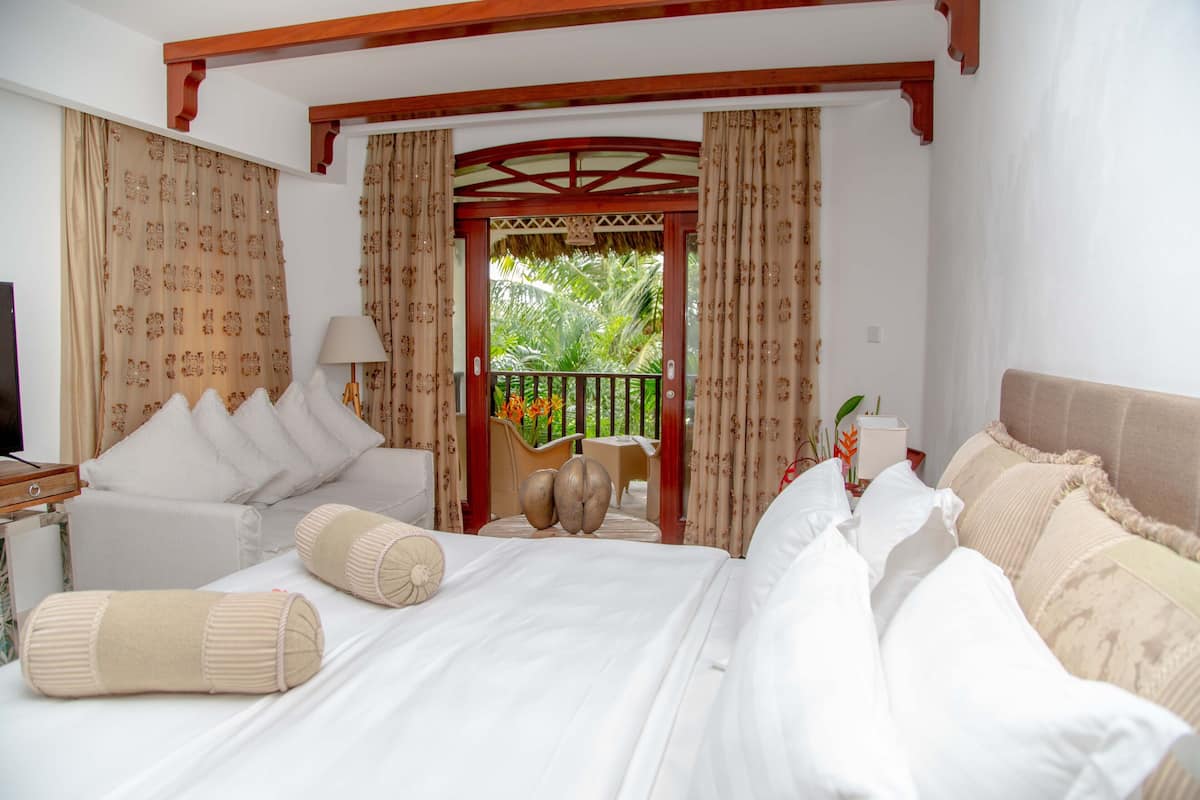 A hotel room with a large bed, two bedside tables, a chair, and a balcony with a view of greenery.