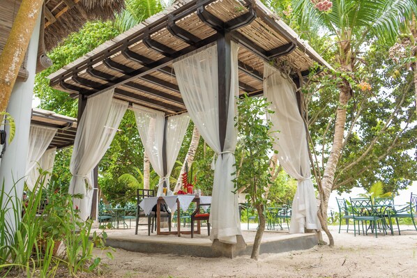 Un espacio de estar techado al aire libre con mesas y sillas, rodeado de follaje y árboles.