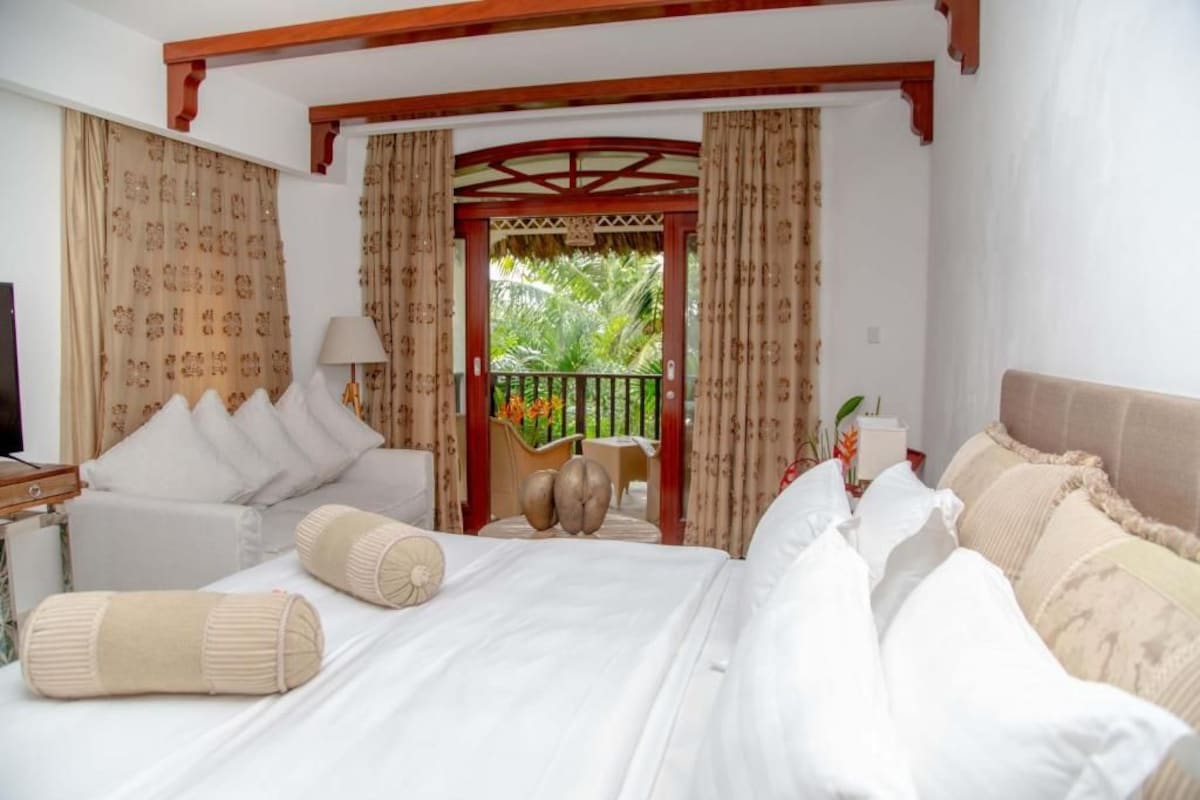 A hotel room with a large bed, a sofa, and a balcony with a view of greenery.