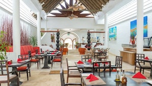 A spacious dining area with high ceilings, wooden beams, and a central open space. Tables are set with red napkins and have a number '6' on them. - Village du Pecheur (Praslin Island)