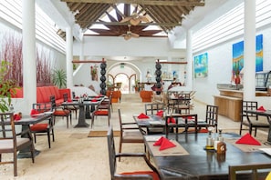 Une salle à manger spacieuse avec de hauts plafonds, des poutres en bois et un espace central ouvert. Les tables sont dressées avec des serviettes rouges et portent le numéro « 6 ».