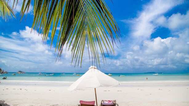 A beach with two sun loungers under a white umbrella, clear blue sky, and calm turquoise sea. - Village du Pecheur (Praslin Island)