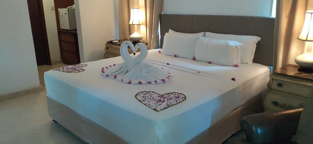 Una habitación de hotel con una cama bien hecha, una decoración con forma de corazón sobre la cama y una mesita de noche con lámpara.