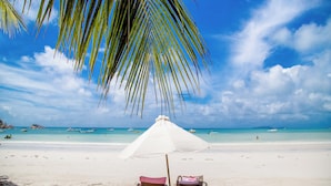 A beach with two sun loungers under a white umbrella, clear blue sky, and calm turquoise sea. - Village du Pecheur (Praslin Island)