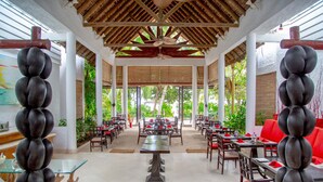 A spacious outdoor dining area with multiple tables and chairs, a central glass-top table, and a ceiling fan. - Village du Pecheur (Praslin Island)
