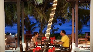 A couple dining at an outdoor table with a view of palm trees and the ocean at dusk. - Village du Pecheur (Praslin Island)