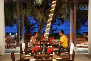 Um casal jantando em mesa ao ar livre, com vista para palmeiras e oceano ao entardecer.