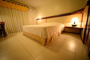 Een hotelkamer met een bed, een nachtkastje met een lamp, een stoel en een raam met jaloezieën.