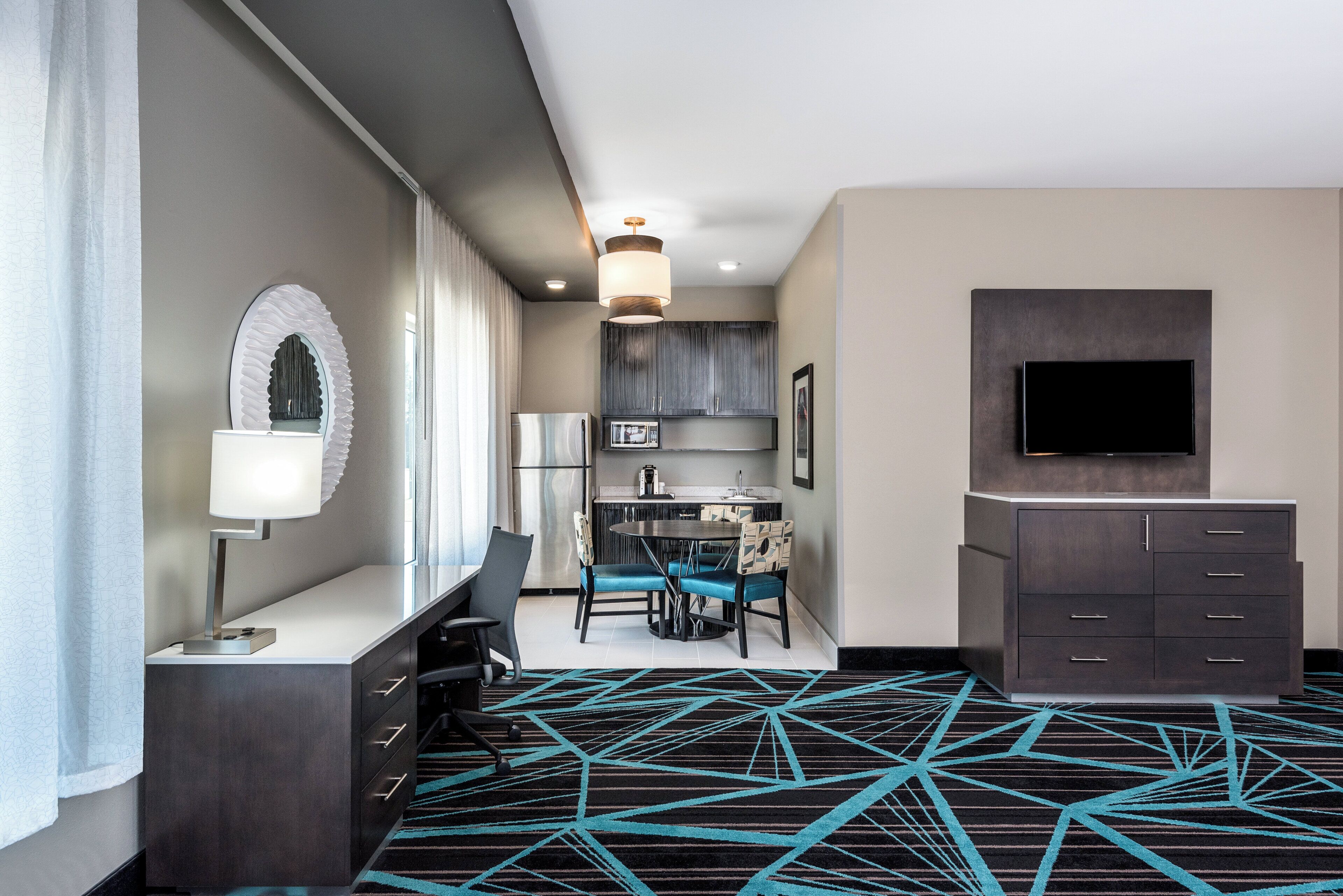 Une chambre d’hôtel équipée d’un bureau, d’une chaise, d’une table et de chaises. On y trouve également un téléviseur fixé au mur, un four à micro-ondes et un réfrigérateur. La chambre est dotée d’une moquette à motifs et d’un luminaire au plafond.