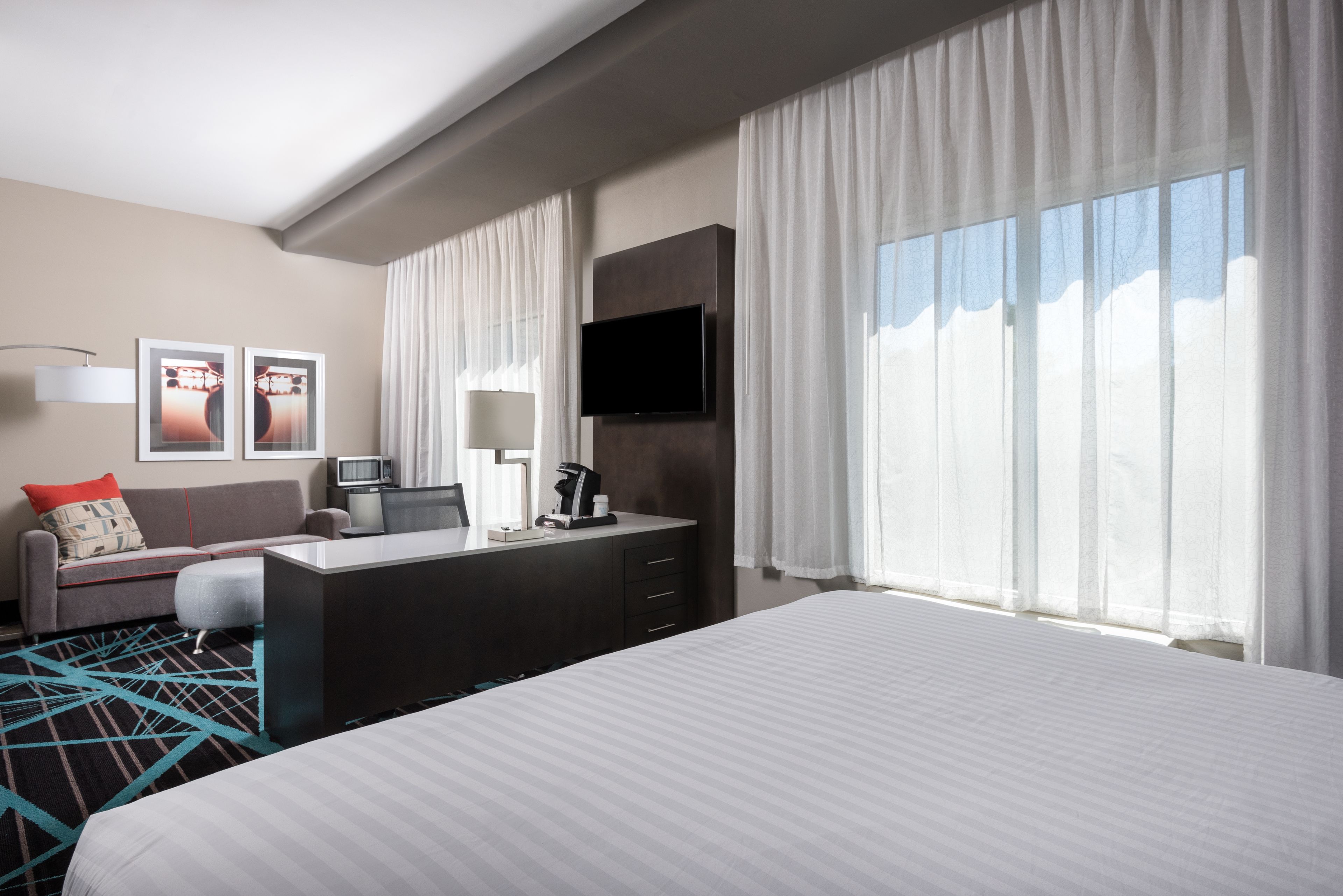 A hotel room with a bed, a desk with a chair, a TV, a coffee maker, and a window with curtains.