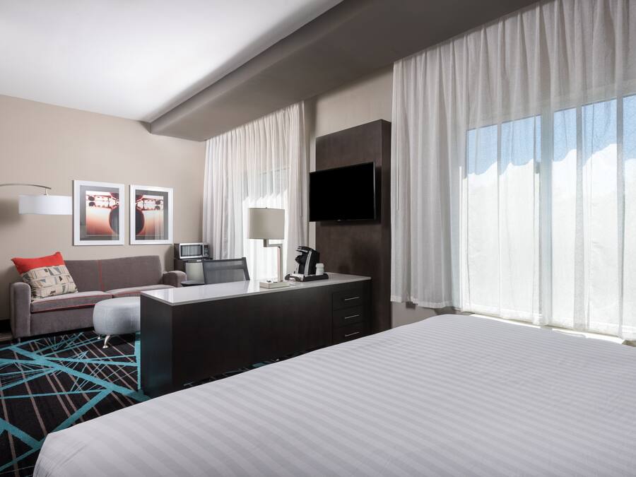 A hotel room with a bed, a desk with a chair, a TV, a coffee maker, and a window with curtains.