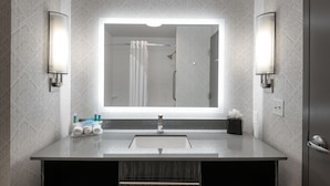 A modern bathroom with a large mirror, two wall-mounted lights, a sink, and a shower area. - Holiday Inn Express & Suites Charlotte Airport by IHG (Charlotte)