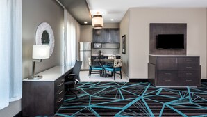 A hotel room with a desk, chair, dining table, and chairs; a television mounted on the wall; a ceiling light fixture; and a patterned carpet. - Holiday Inn Express & Suites Charlotte Airport by IHG (Charlotte)