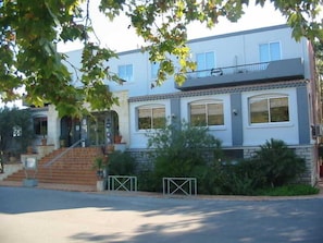 Un edificio residencial de dos pisos con entrada principal, balcón y un camino de acceso con portón.