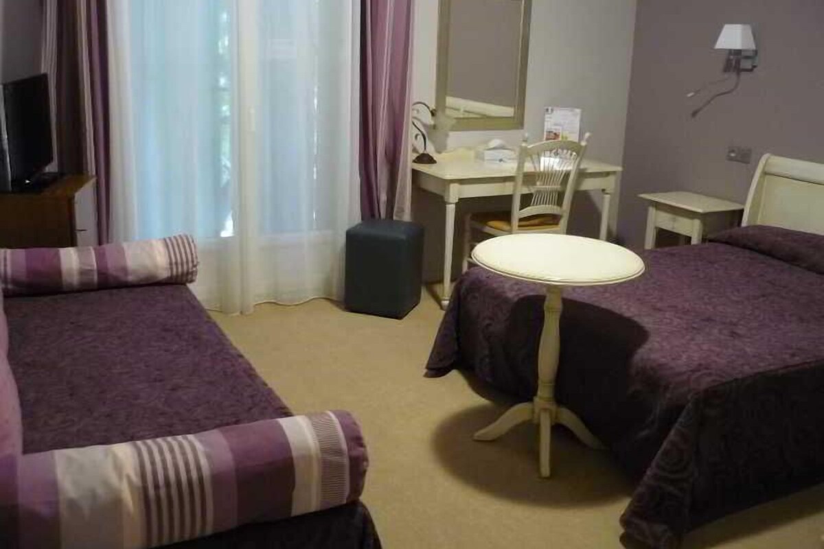 Habitación de hotel con dos camas, un escritorio, una silla, una mesita, una lámpara y una ventana con cortinas.