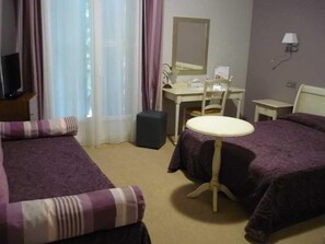 Habitación de hotel con dos camas, un escritorio, una silla, una mesita, una lámpara y una ventana con cortinas.