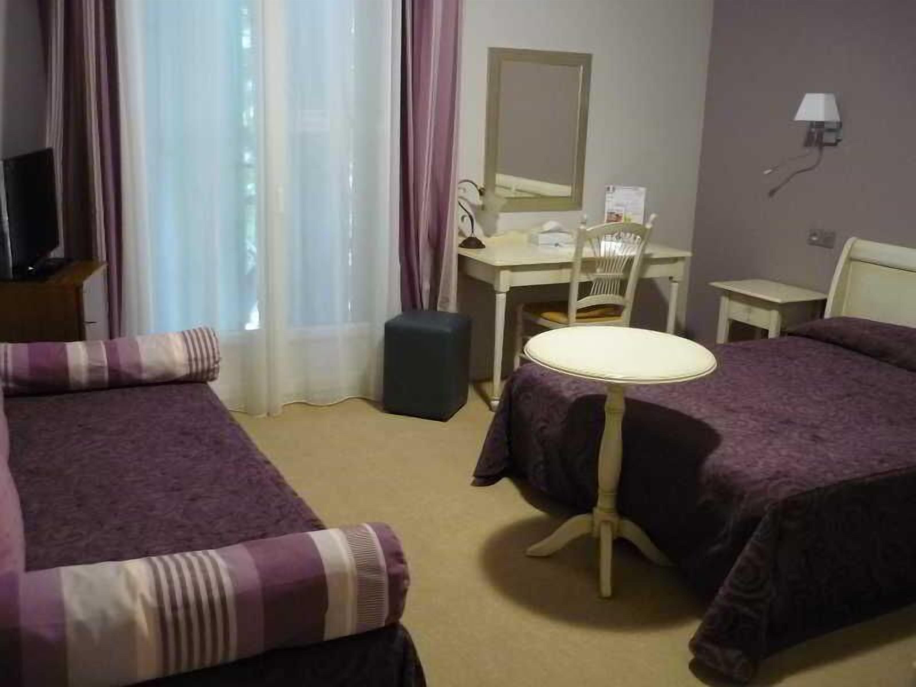 Habitación de hotel con dos camas, un escritorio, una silla, una mesita, una lámpara y una ventana con cortinas.