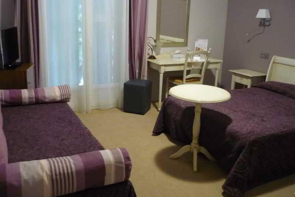 A hotel room with two beds, a desk, a chair, a small table, a lamp, and a window with curtains.
