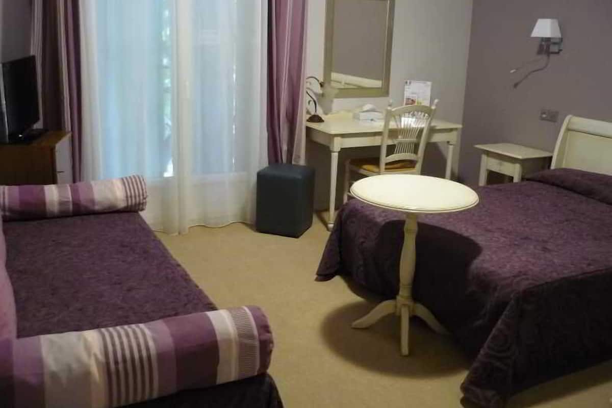 Habitación de hotel con dos camas, un escritorio, una silla, una mesita, una lámpara y una ventana con cortinas.