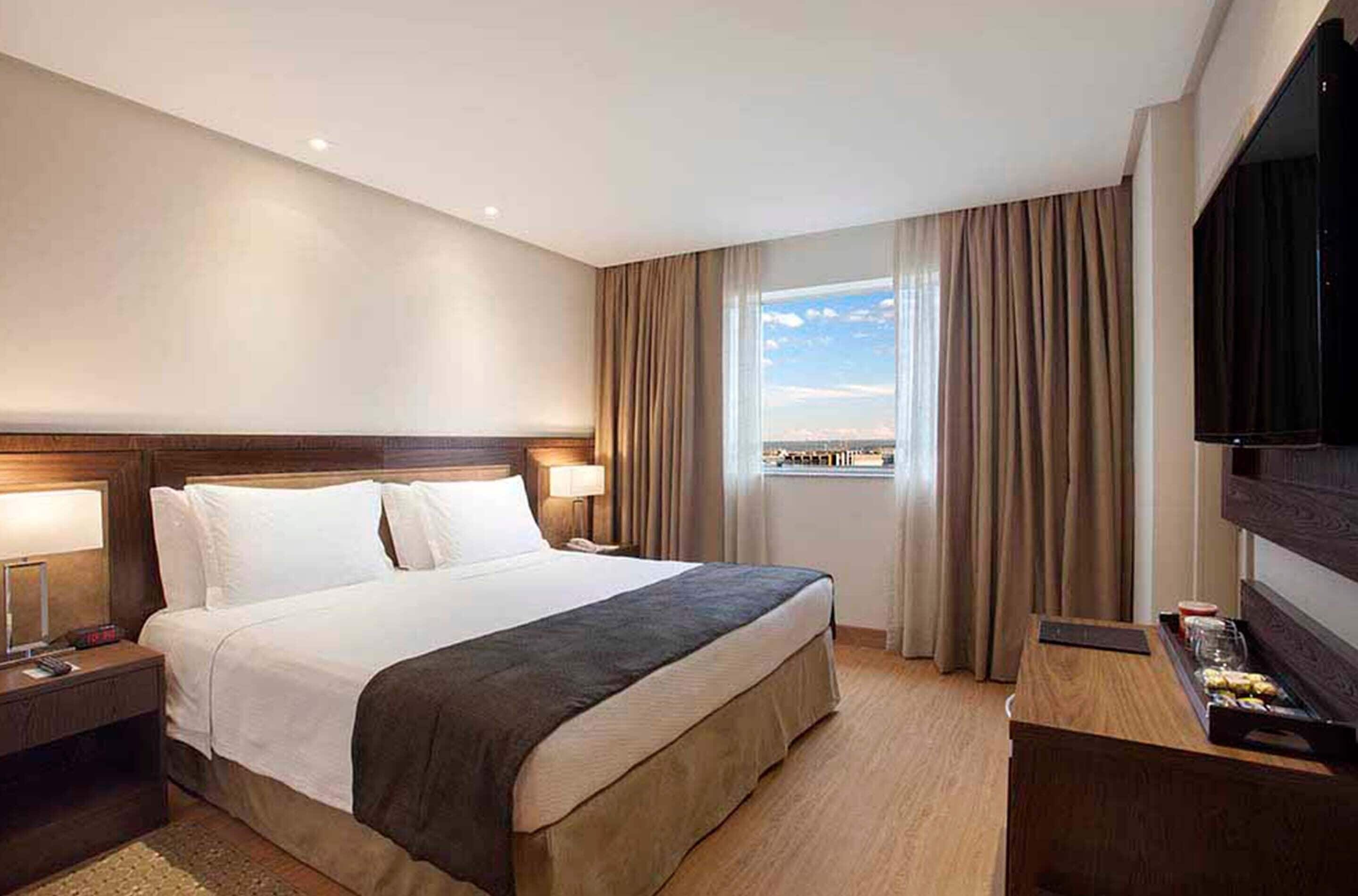 Habitación de hotel con una cama grande, televisión, escritorio con una bandeja de aperitivos y ventana con cortinas.