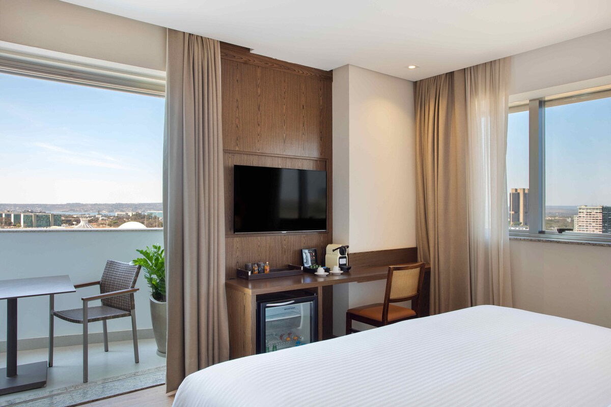 Ein Hotelzimmer mit Bett, Schreibtisch mit Fernseher, Kaffeemaschine, Minikühlschrank, Stuhl und einer Zimmerpflanze.