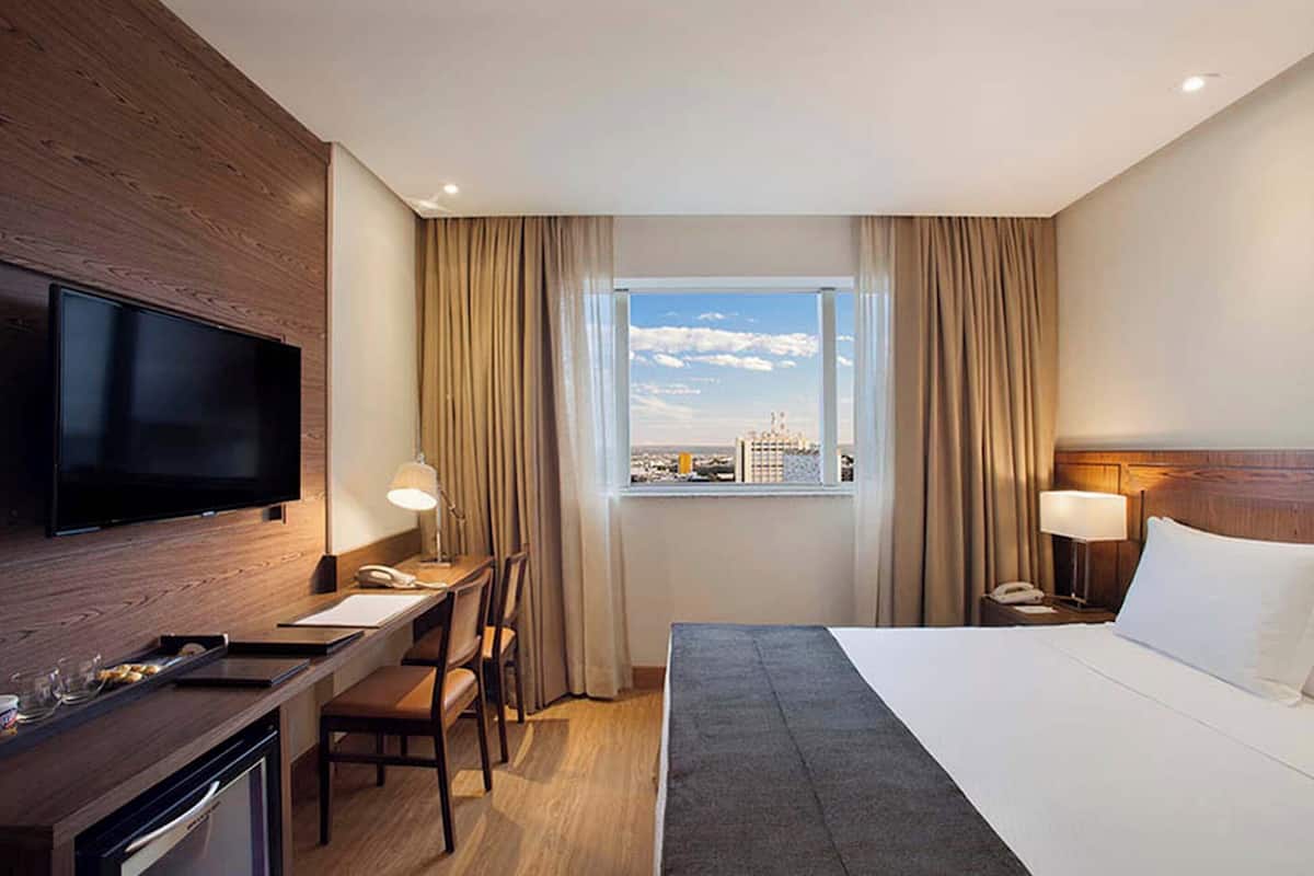 Habitación de hotel con cama, escritorio, silla, televisor y ventana con vistas a la ciudad.