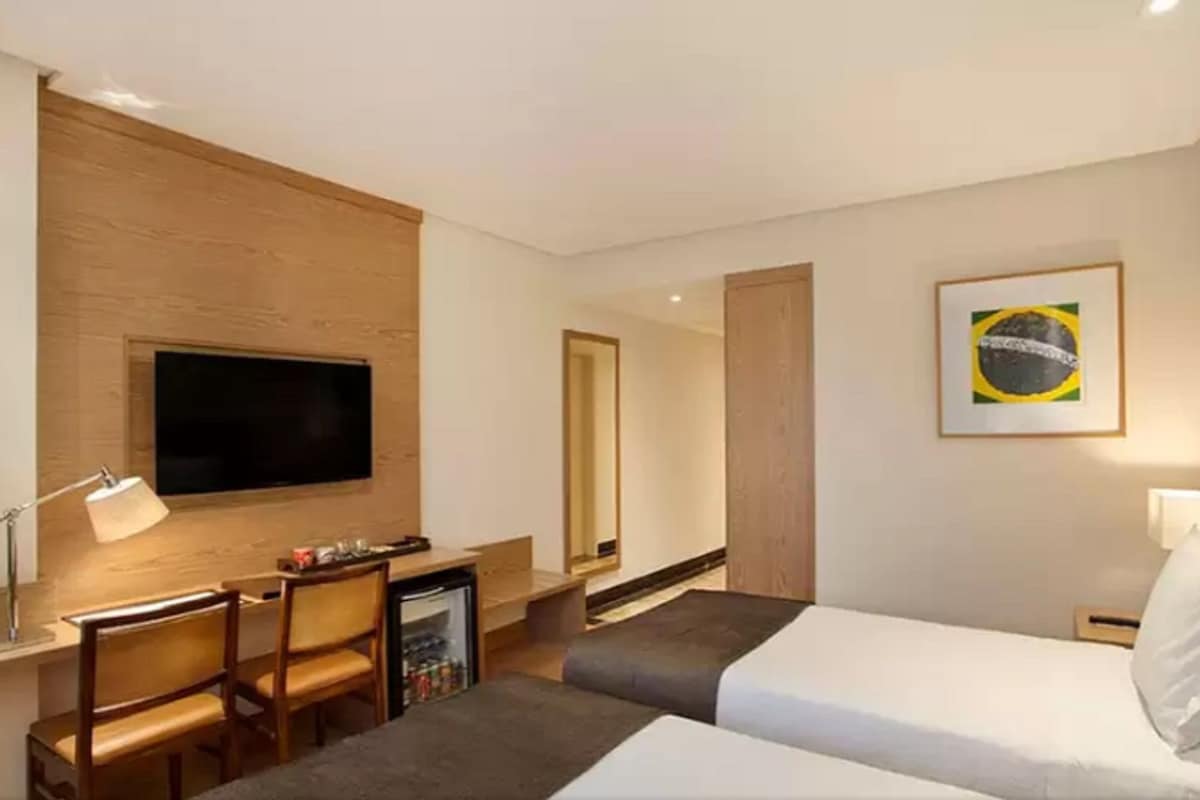 Une chambre d’hôtel comprenant un lit, un bureau avec une chaise, un téléviseur fixé au mur, un petit réfrigérateur et un tableau représentant le drapeau brésilien.