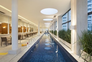 Uma piscina interior extensa com piso de azulejos, rodeada por áreas de estar e grandes janelas.