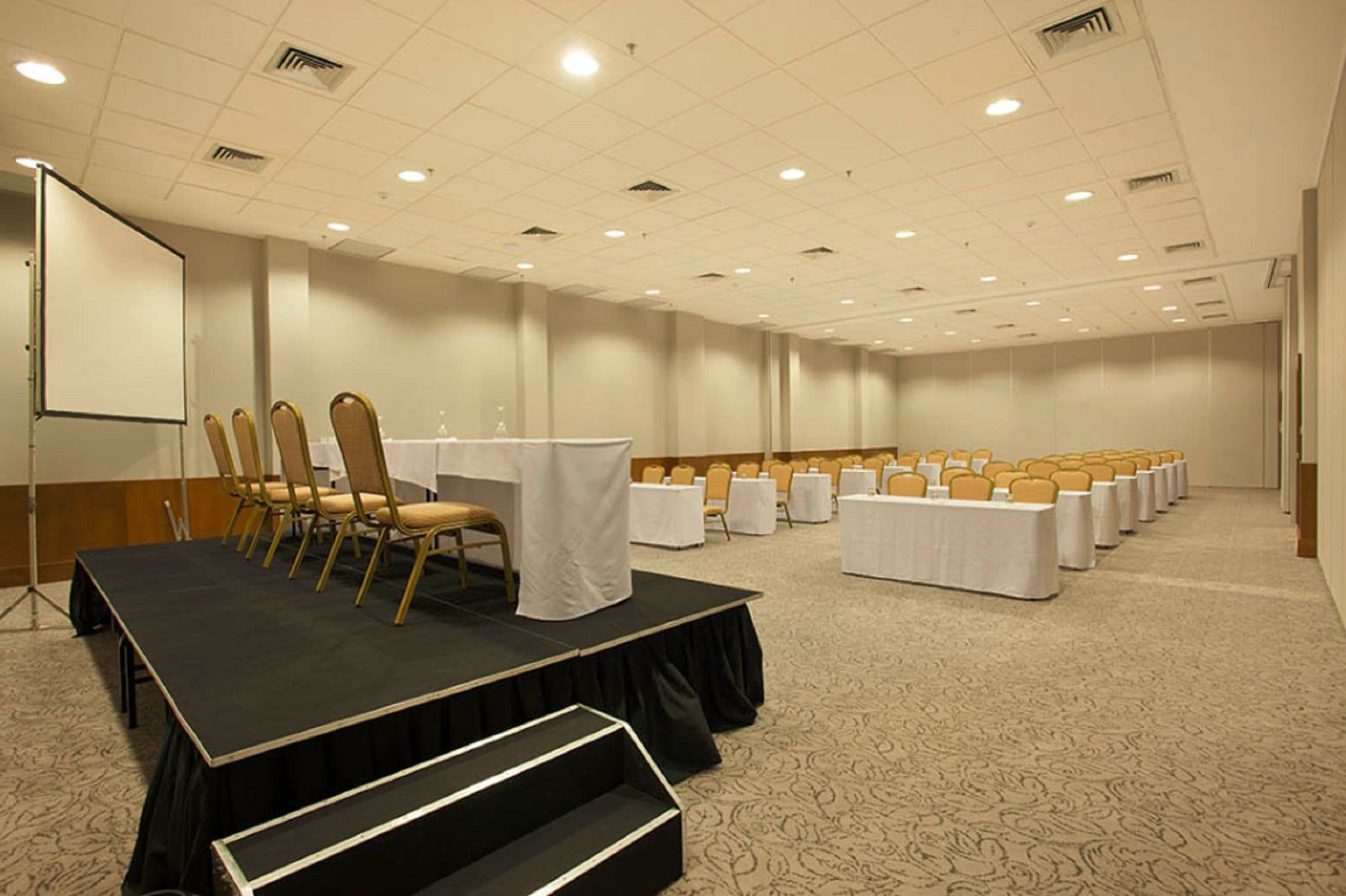 Sala de conferências com palco, mesas e cadeiras organizadas em fileiras.